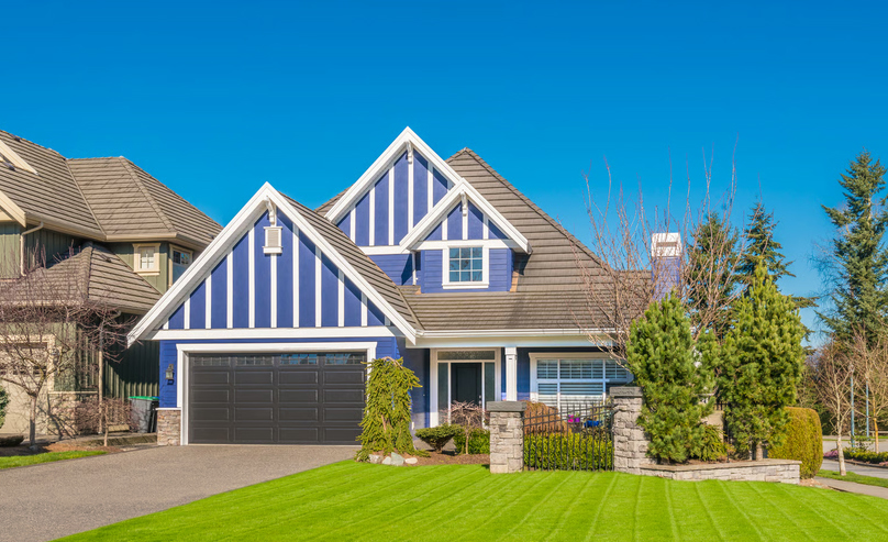 Two-story blue craftsman house with white trim, dark roof, front garage, manicured green lawn, and clear blue sky